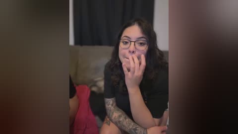 Video of a tattooed woman with curly dark hair, glasses, and black top, covering her mouth in surprise, sitting on a couch with a pink blanket in a dimly lit room.