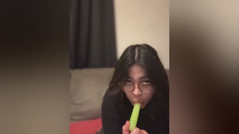 Video of an Asian woman with long black hair, wearing glasses, eating a green banana in a dimly lit room with beige walls and red carpet.