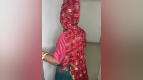 Video of a person wearing a red and white striped shirt, a red and white patterned sari, and a yellow bracelet, standing in a doorway with blurred background.