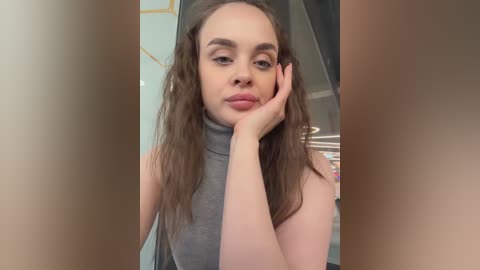 Video of a fair-skinned, long-haired, brunette woman with a slim build and medium-sized breasts, wearing a sleeveless grey turtleneck, resting her chin on her hand, in an indoor setting with a blurred background.