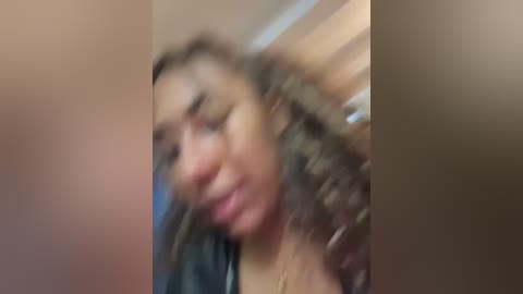 Media: A blurred video of a young woman with curly hair, wearing a black top, with a background of a wooden door and beige wall.