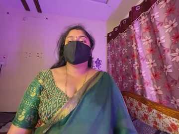Media: Video of a South Asian woman in a green sari with a floral border, wearing a black mask, sitting in a room with floral curtains and a wooden headboard.