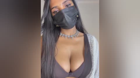 Media: Video of a dark-skinned woman with long black hair, wearing a black face mask, black bra, and a white cardigan, set against a blurred beige background.