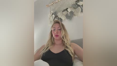 Video of a blonde woman with fair skin, wearing a black tank top, sitting on a bed with white sheets. She has long, wavy hair and a neutral expression. The background features a hanging woven wall decoration.