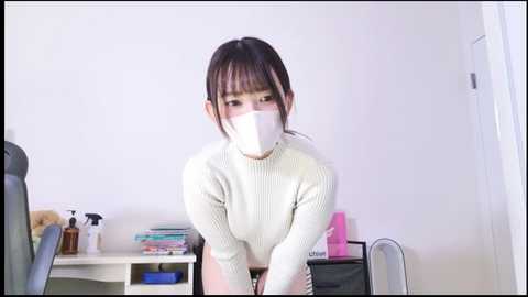 Media: A video of an Asian woman with straight black hair and bangs, wearing a white ribbed sweater, white mask, and black pants, leaning over a desk in a tidy office.