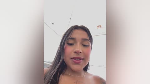 Video of a young, Latina woman with long, straight black hair, fair skin, and light makeup. She has a neutral expression and is topless, showing a small tattoo on her chest. The background is a white room with a ceiling fan.