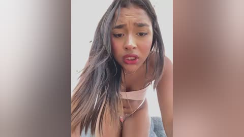 A video featuring a young woman with long, straight brown hair, wearing a light pink bra and panties, leaning forward with a surprised expression, blurred background.