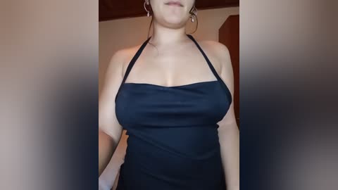 Video of a fair-skinned woman with shoulder-length brown hair, wearing a black halter top and tight black pants, standing indoors against a beige wall.