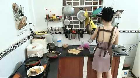 Video of an Asian woman with dark hair in a bun, cooking in a small, cluttered kitchen with white walls and black countertops. She wears a brown apron over a sleeveless top. Pots, pans, and utensils hang from a metal rack above the stove.