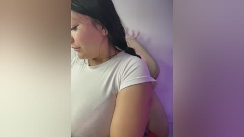 Video of a young Asian woman with straight black hair, wearing a white T-shirt, pressed against a white wall, with a purple and orange gradient background.