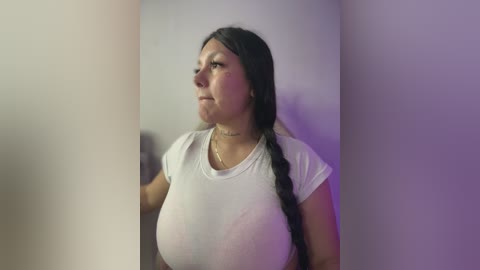 Video of a Latina woman with long dark hair, large breasts, wearing a tight white t-shirt, standing against a plain white wall with purple lighting.