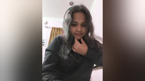 A video of a young woman with long, dark hair, wearing a black sweater, standing indoors with a blurred background. She is touching her throat and smiling slightly.