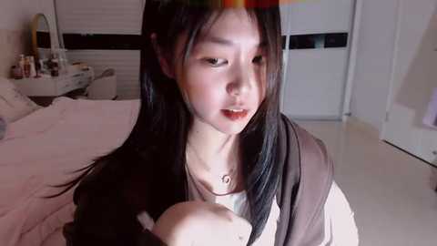 Media: Video of an Asian woman with long black hair, fair skin, and a slender build, wearing a brown jacket and white top, sitting on a bed in a dimly lit room with white walls and closed blinds.