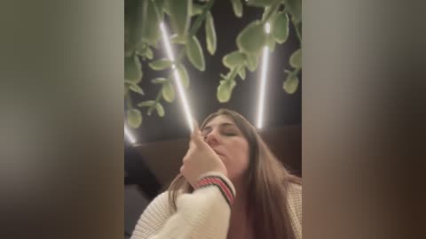Video of a fair-skinned woman with long brown hair, wearing a cream sweater, smoking a cigarette, surrounded by hanging green plants under fluorescent lighting.