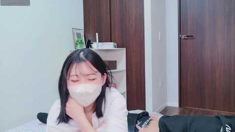 Video of an Asian woman with long black hair and a white mask, lying on a bed, looking distressed. Background features wooden wardrobe and a small table with items.