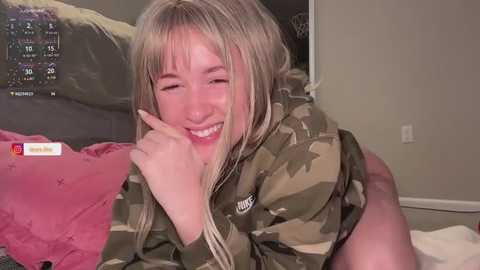 A video of a blonde woman with straight hair, wearing a camo jacket, smiling, on a bed with pink pillows.