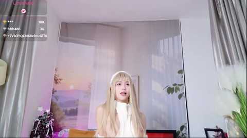 A video of a young, fair-skinned woman with long, straight blonde hair, wearing a white turtleneck, standing in a brightly lit room with a mural of a sunset and potted plants.