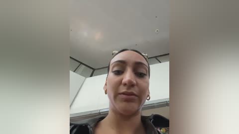 Video of a young woman with light brown skin, dark hair in buns, wearing hoop earrings, and a black hoodie. She stands indoors in a modern room with white walls and recessed lighting.