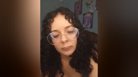 Media: Video of a young woman with curly black hair, fair skin, wearing round glasses, and a neutral expression, standing against a wall adorned with colorful, abstract art.