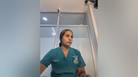Video of a young Latina nurse with light brown skin and brown hair tied back, wearing teal scrubs with an American Heart Association logo, standing in a well-lit hospital room with large windows.