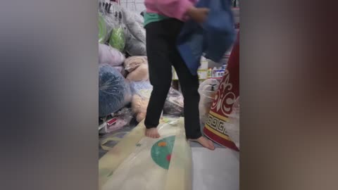Video of a barefoot person, wearing a pink top and black pants, standing amidst a cluttered room filled with plastic bags, toys, and a red box. The scene appears cramped and disorganized.