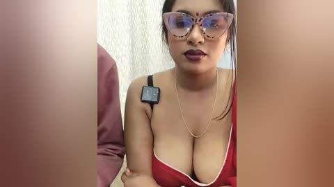 Media: Video of a voluptuous, light-skinned woman with long black hair and large breasts, wearing red lingerie and large, transparent, cat-eye glasses. She's seated indoors, with a blurred background.