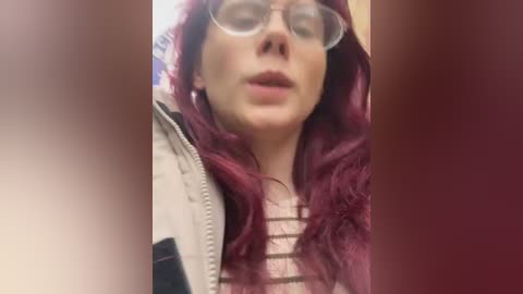 Media: Video of a fair-skinned woman with long, wavy, dark red hair, wearing round glasses and a striped shirt, standing indoors.