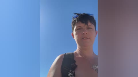 A video of a woman with short dark hair, wearing a black sleeveless top with a silver necklace, standing in front of a white pillar against a clear blue sky.