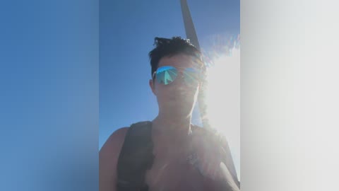 Video of a shirtless man with short dark hair, wearing reflective sunglasses, standing against a bright blue sky with a sun flare, holding a cigarette.