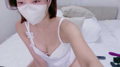 A video of an Asian woman with fair skin and medium-length brown hair, wearing a white lace bra and face mask, sitting on a white bed, with a remote control and a purple vibrator on the bed.