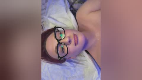 Video of a fair-skinned woman with red hair, wearing black-rimmed glasses, lying on a bed with white sheets, partially nude, with a blurred figure in the foreground.