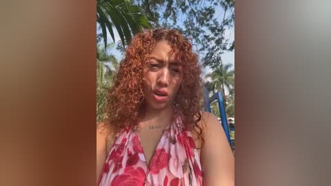 Video of a young woman with curly, reddish-brown hair, wearing a red and white floral halter top, standing outdoors with lush green palm trees and blue sky in the background.