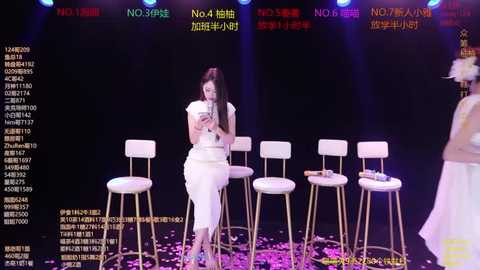 Video of a female TV host in white dress, seated on a stool, surrounded by white chairs, in a brightly lit studio with colorful graphics and text in Chinese.