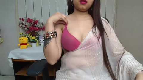 Video of a woman with long black hair, wearing a pink bra, and a white sheer sari, sitting on a chair, against a background of a table with toys and flowers.