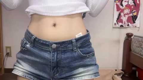 Video of a person wearing a white crop top and blue denim shorts with a pocket. Background features a bed, a wall poster, and a power outlet.