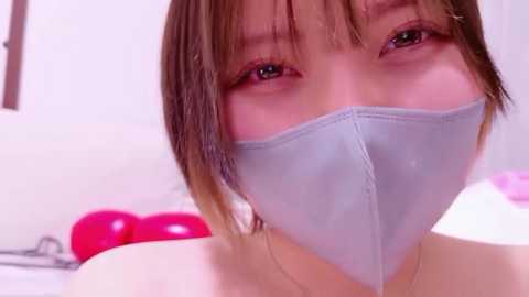Video of an Asian woman with short brown hair, wearing a light blue surgical mask, and a white top, in a brightly lit room with pink objects in the background.