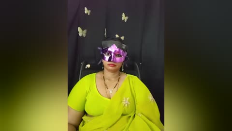 Media: Video of a woman with medium brown skin, wearing a bright yellow sari with floral embroidery, a purple mask, and butterfly earrings, seated against a black backdrop with butterflies.