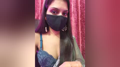 Video of a South Asian woman with long, straight black hair, wearing a blue lace bra, black mask, and dangling earrings, against a red velvet backdrop.