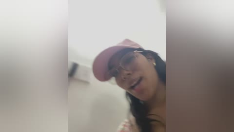 Media: A blurry photo of a smiling woman with long dark hair, wearing a pink cap and glasses, taken in a dimly lit bathroom with white walls and a mirror.