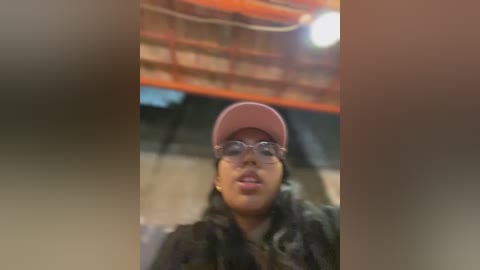 Media: A video of a young woman with medium skin tone, wearing a pink cap and glasses, standing indoors in a gym with red and orange walls.