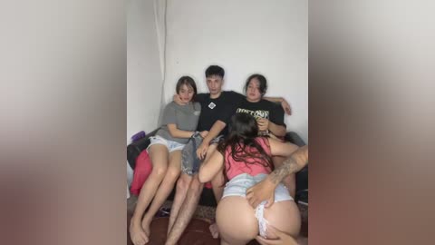 A video captures four young adults sitting in a dimly lit room. They are dressed casually, with one person partially undressed, revealing a tattooed arm. The background is plain, focusing on the group.