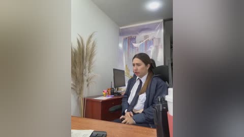 Media: Video of a woman in a navy suit seated at a wooden desk in a minimalist office with beige walls, a tall potted plant, and a computer monitor.