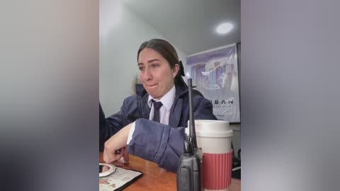 Media: Video of a woman with long brown hair in a dark suit, holding a pen and a white coffee cup with a red stripe, seated at a wooden desk in a brightly lit office with a Bank of America poster on the wall.