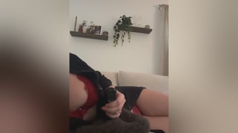 Media: Video of a woman in red lingerie and a black dress, holding a black gun, seated on a beige couch. Background includes a white wall with two floating shelves, potted plants, and a curtain.