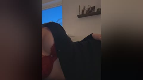 Video of a woman in a black dress with a red bra, bending over. The background shows a white wall with a shelf holding small items, and a window with blue light filtering through.