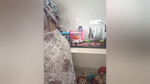 Media: A video of a small, cluttered kitchen featuring a person with a patterned shirt, white tiles, a sink, and various utensils.