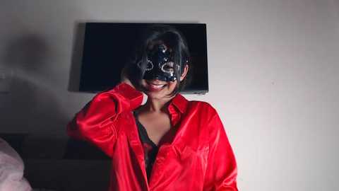 Media: Video of a woman with medium brown skin and straight black hair, wearing a shiny red satin robe and a black gas mask, smiling, in a dimly lit room with a white wall and a flat-screen TV.