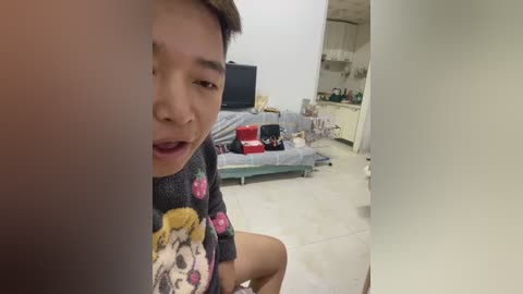 Video of a young Asian man in a messy bedroom with a bed, TV, and toys, wearing a gray t-shirt with cartoon characters.