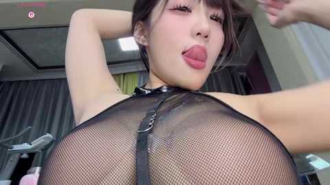 Media: Video of an Asian woman with fair skin, wearing a black fishnet bodysuit, sticking out her tongue, posing seductively in a gym setting.