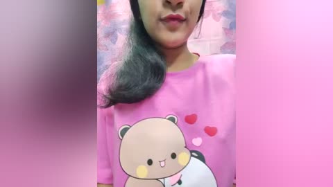 Media: Video of a woman with medium-length black hair, wearing a pink t-shirt with a cartoon bear and hearts, smiling against a pink background.